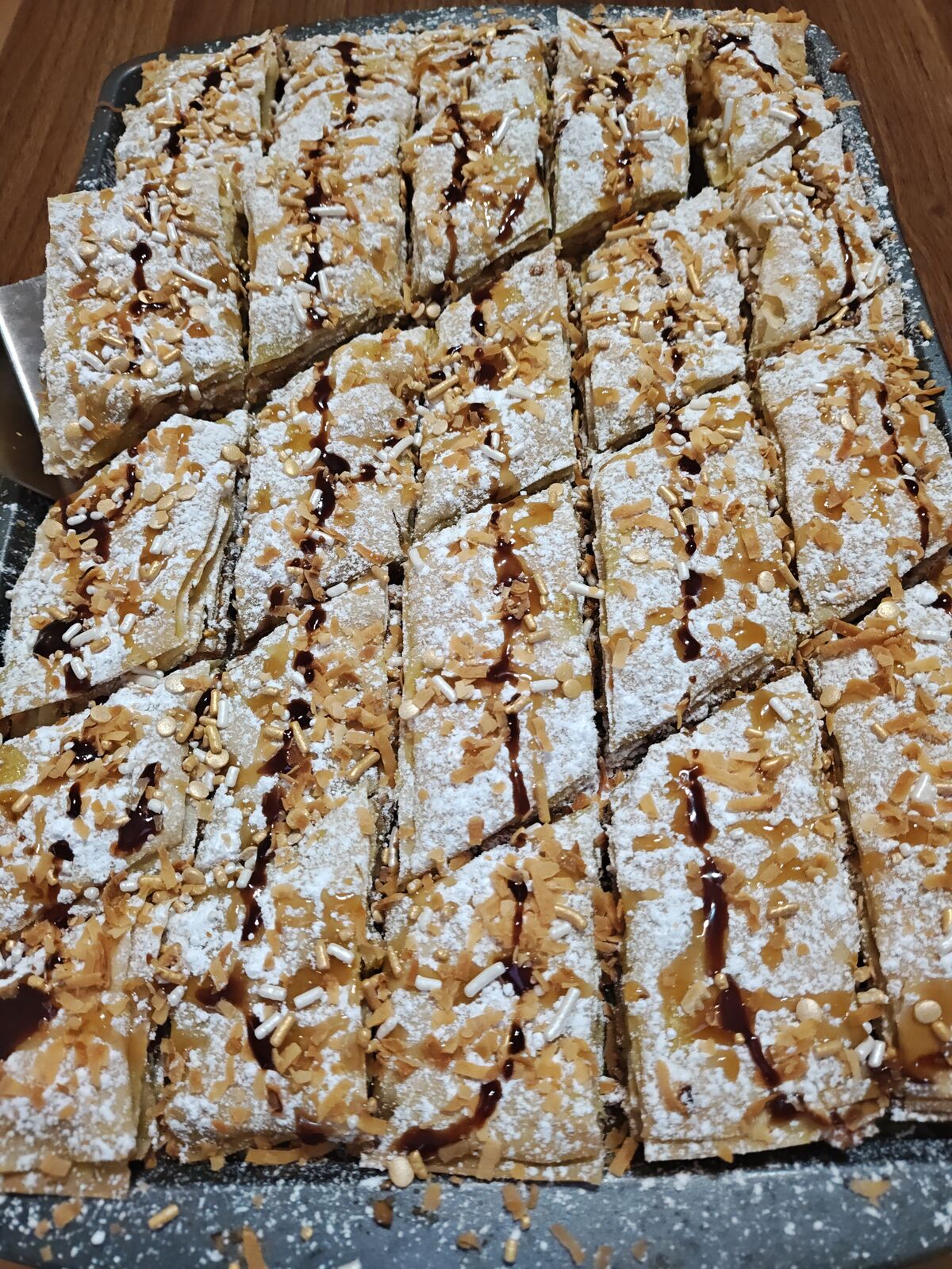 A freshly cut tray of baklava finished with powdered sugar, toasted coconut, gold sprinkles and a chocolate drizzle